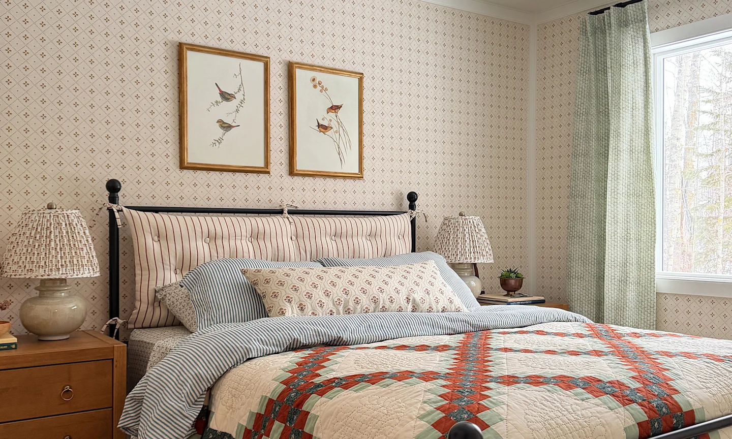 Cabin bedroom with neutral wallpaper, striped bedding in red and blue and green curtains.