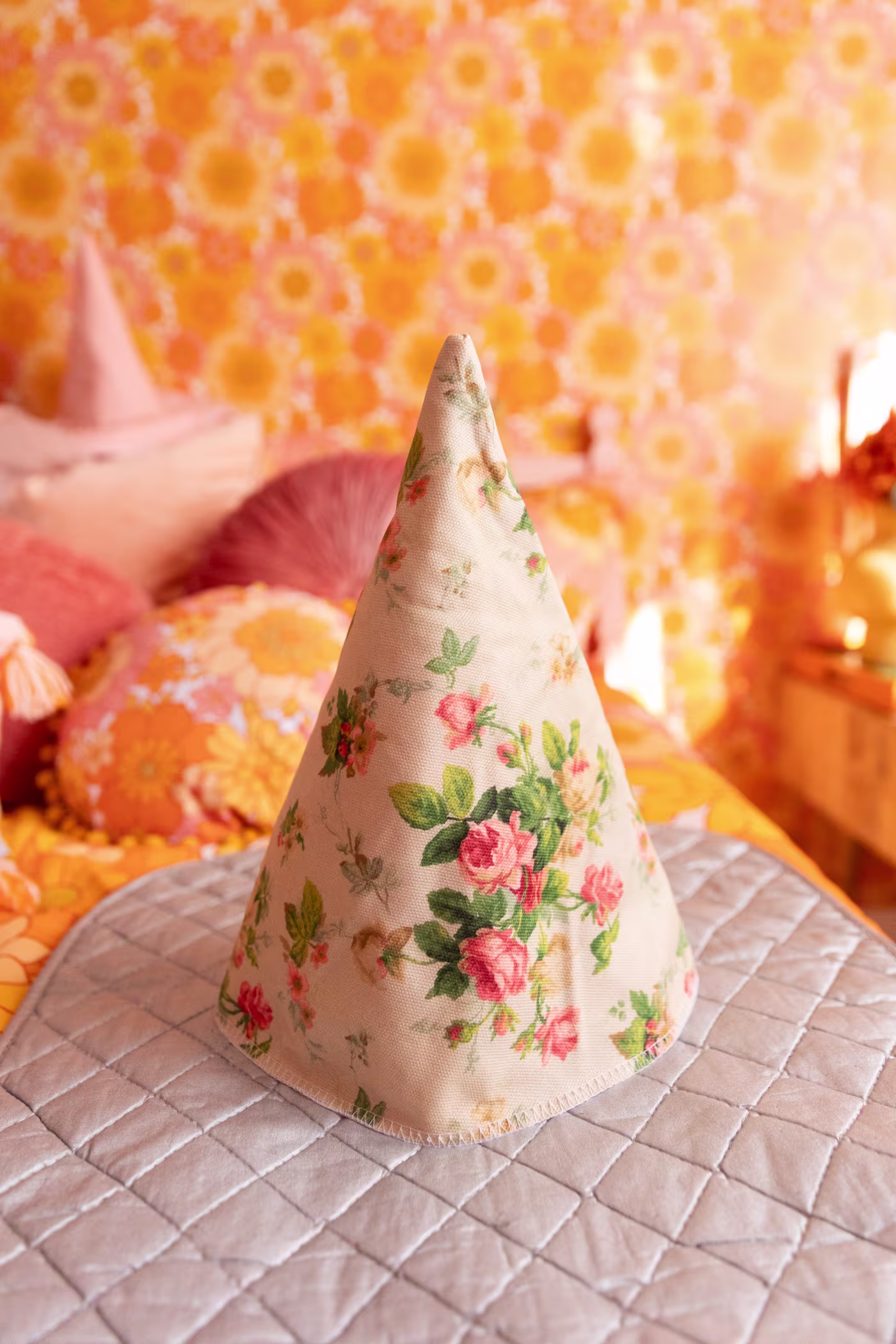 A cream colored cone with pink roses and green leaves sits on a silver ironing mat. The mat is resting on a bed with retro floral bedding. Out of focus, to the left are throw pillows in different shades of pink. Wallpaper, that matches the bedding is on t
