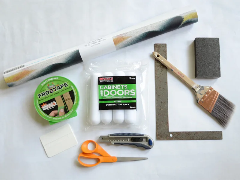 materials to wallpaper a dresser lay on a white background. Materials include a roll of wallpaper, painters tape, a squeegee, scissors, a sanding block, a measuring device and craft knife.