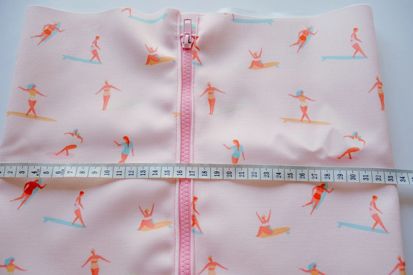 A fabric rectangle featuring a design with small female surfers surfing through a pink background lays on a white surface. A pink zipper has been stitched up the middle. A small white measuring tape has been placed over the fabric.