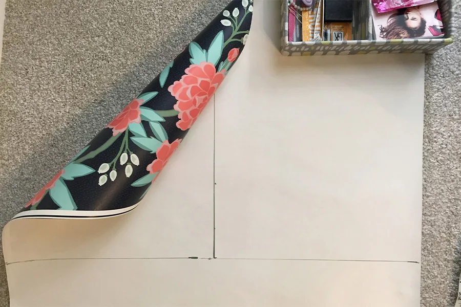 A piece of wallpaper laying design side down on carpet. Lines indicating measurements are on the wrong side. A small box in the corner keeps the wallpaper from rolling up.