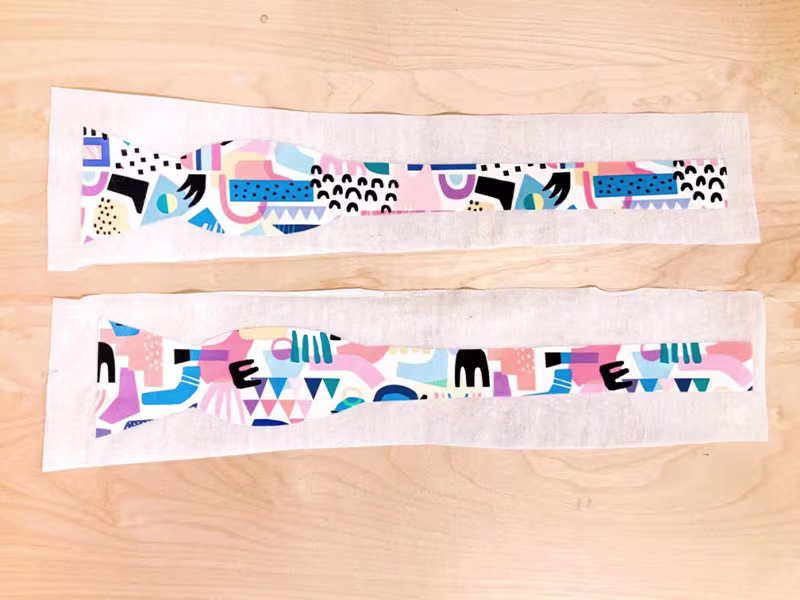Two bow tie pieces lay design side up on two pieces of interfacing on a wooden surface.