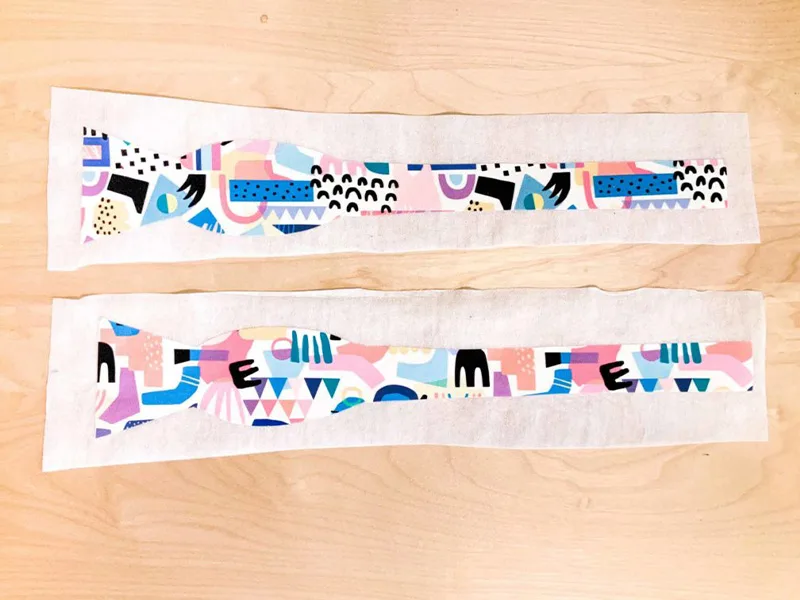 Two bow tie pieces lay design side up on two pieces of interfacing on a wooden surface.