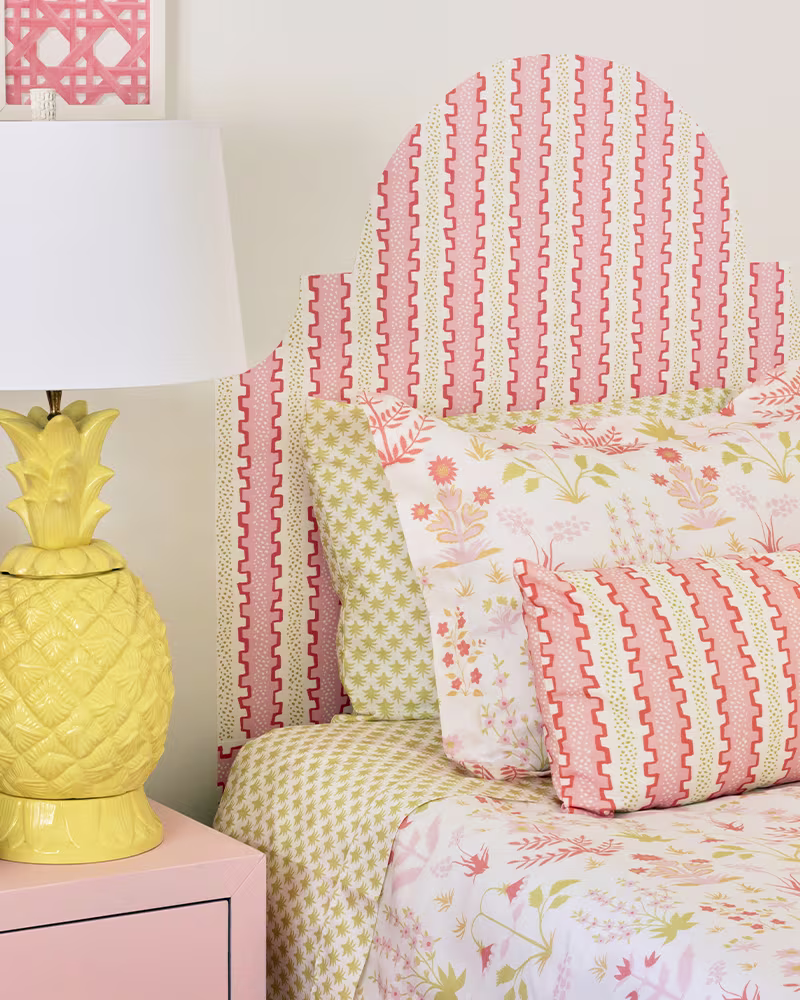 A close up of the finished headboard and the top of the made-up bed, along with a piece of artwork to the top left. A yellow lamp has been placed on a pink bedside table to the left of the bed. The designs on the art, bedding and headboard are light pink
