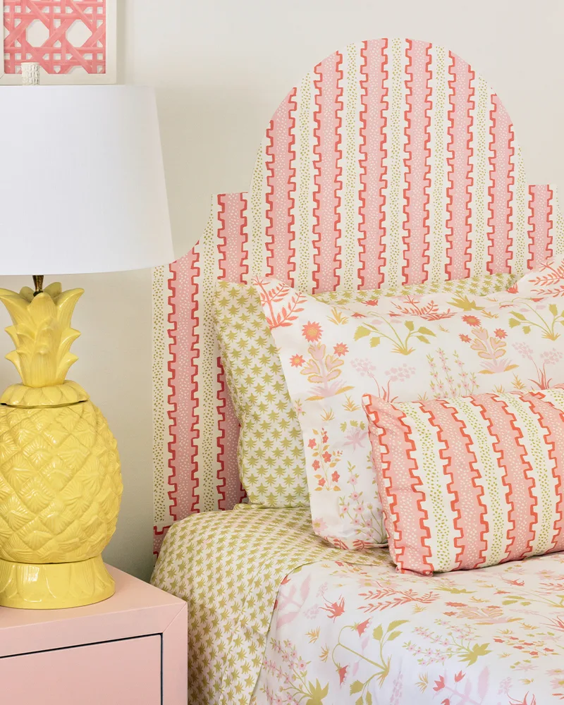 A close up of the finished headboard and the top of the made-up bed, along with a piece of artwork to the top left. A yellow lamp has been placed on a pink bedside table to the left of the bed. The designs on the art, bedding and headboard are light pink