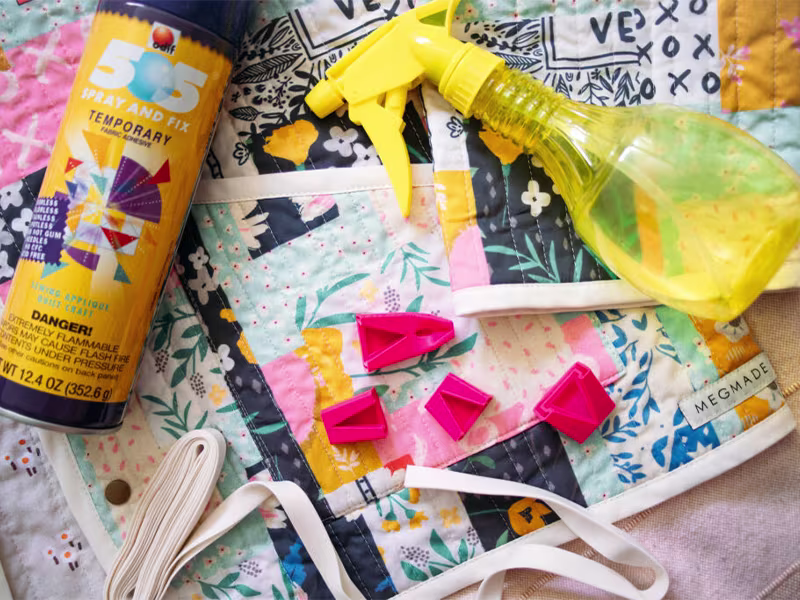 Materials to make quilted jacket are laid out on a blue cheater quilt quilted jacket. Including a hot pink 3D printed bias tape maker, some temporary Spray and Fix, white bias tape and a yellow spray bottle to fill with homemade spray starch.