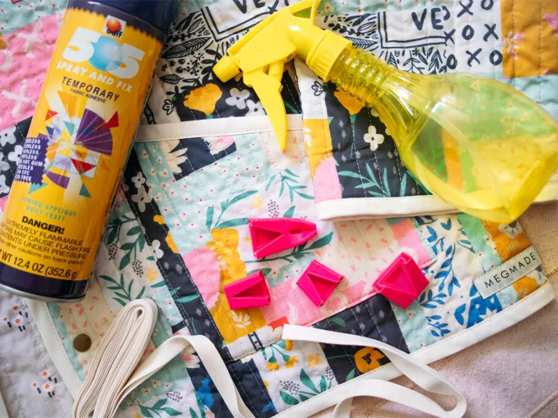 Materials to make quilted jacket are laid out on a blue cheater quilt quilted jacket. Including a hot pink 3D printed bias tape maker, some temporary Spray and Fix, white bias tape and a yellow spray bottle to fill with homemade spray starch.