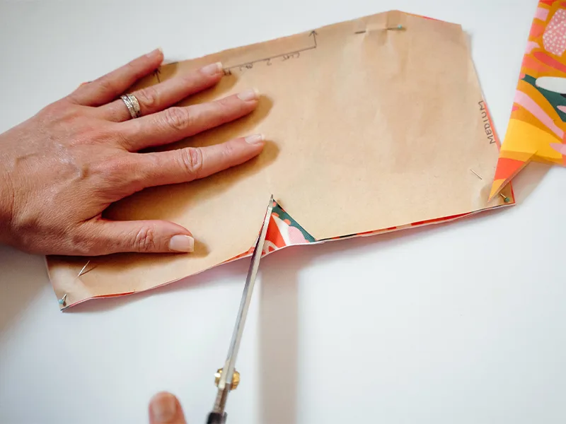 Cutting out the bowl cozy paper pattern that has been pinned and placed on the fabric fold. It is being cut out along the wide V-shaped notch along the one of the pattern’s long sides. The fabric design features bright flowers in pinks, reds, yellows and 