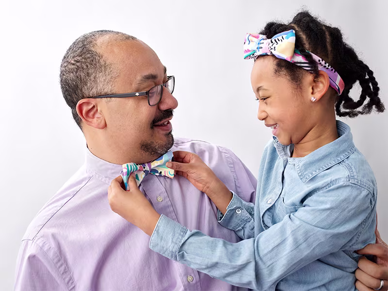 A little girl holding a bowtie that a man is wearing. She's wearing a matching headband.