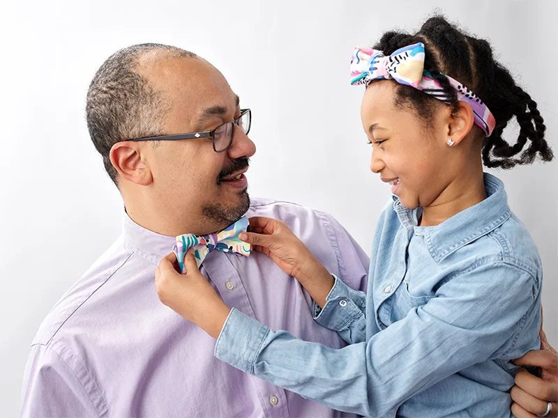 A little girl holding a bowtie that a man is wearing. She's wearing a matching headband.