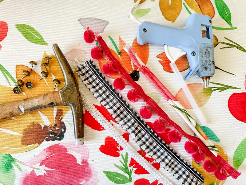 Tacks, a tack hammer, gimp, ribbon, pompom trim and double welt cord and a hot glue gun rest on a floral background.
