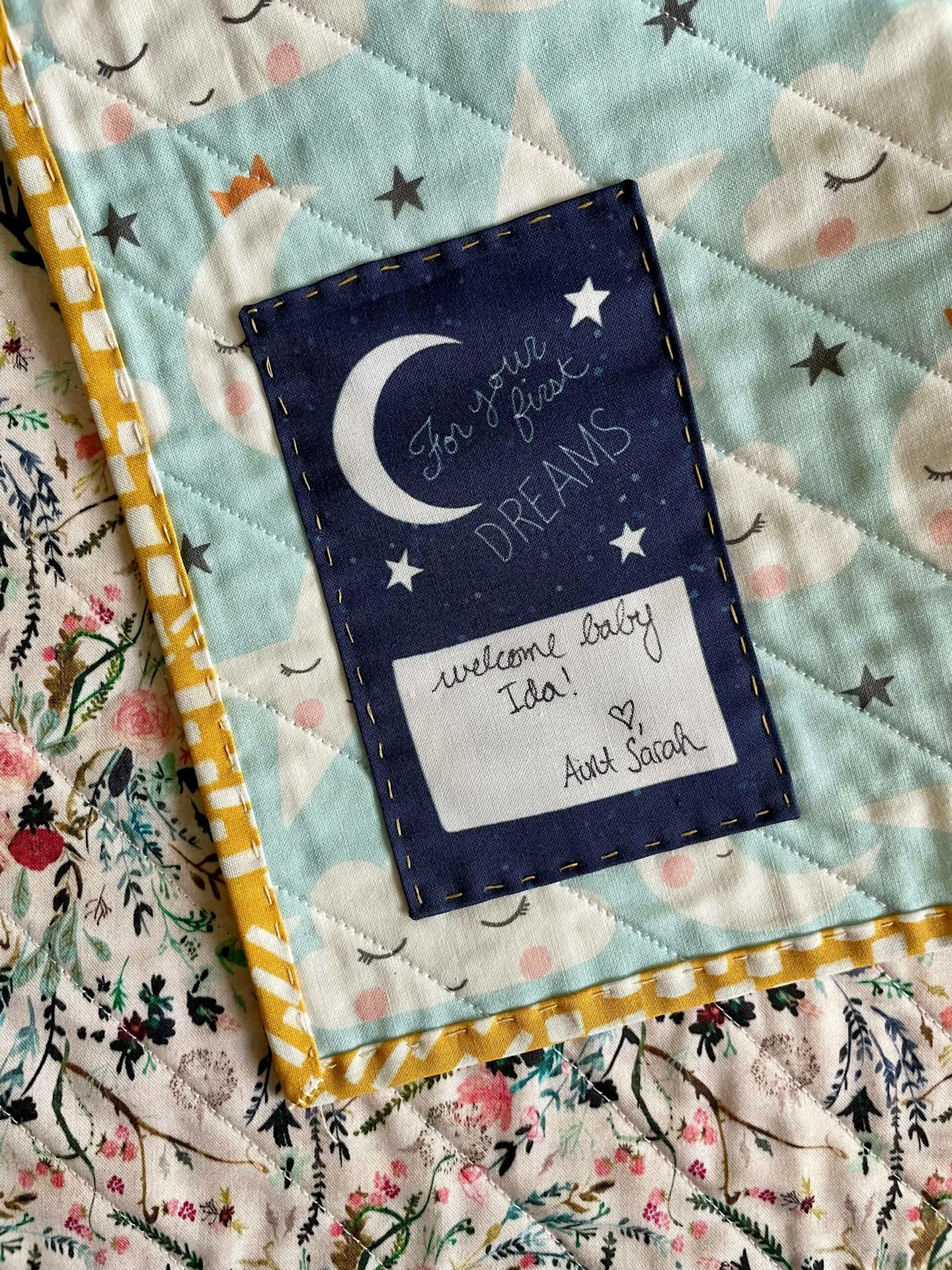 The top of a quilt label sewn to the bottom left corner of a quilt has a navy box with a white crescent moon and small white stars that says “For your first dreams” in a white cursive font. In a white box underneath the navy box, someone has written “Welc