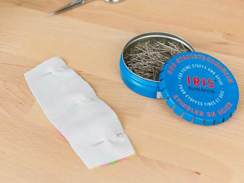 A rectangle piece of fabric lays design side down with two pins and either horizontal end. The container holding the pins is nearby.