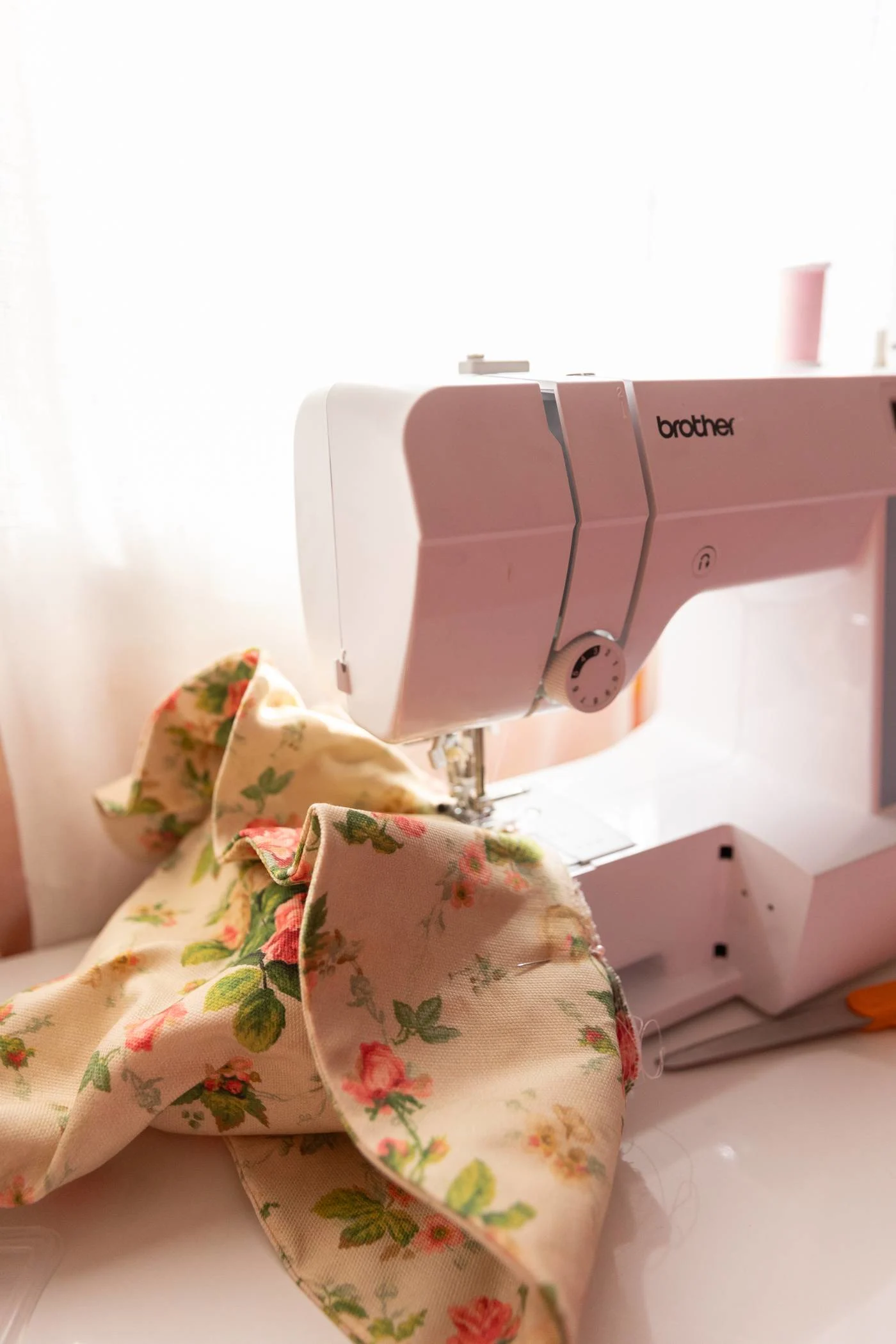 A sewing machine stitches the brim to the cone of the witch hat on a white desk. A pair of scissors are beside the machine. A sheer white curtain filters light from a nearby window.