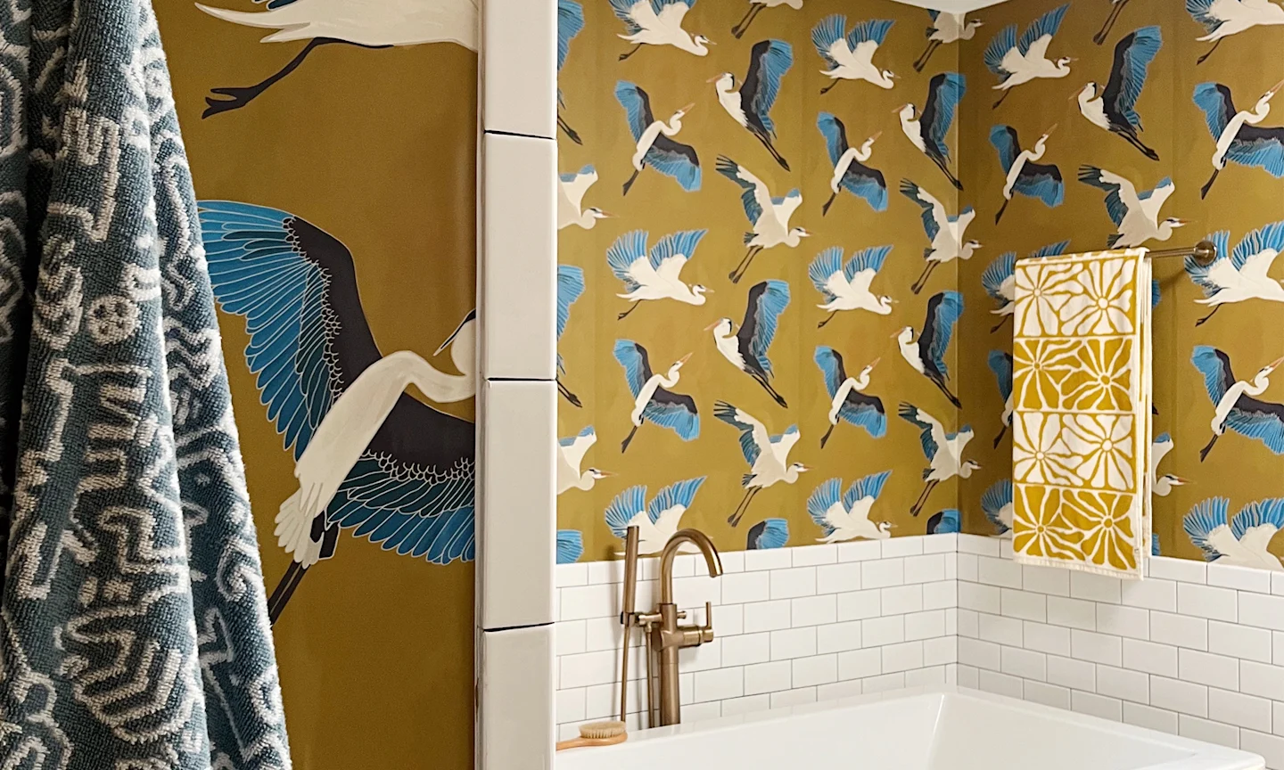 Bathroom with gold flying herons wallpaper above a bathtub.