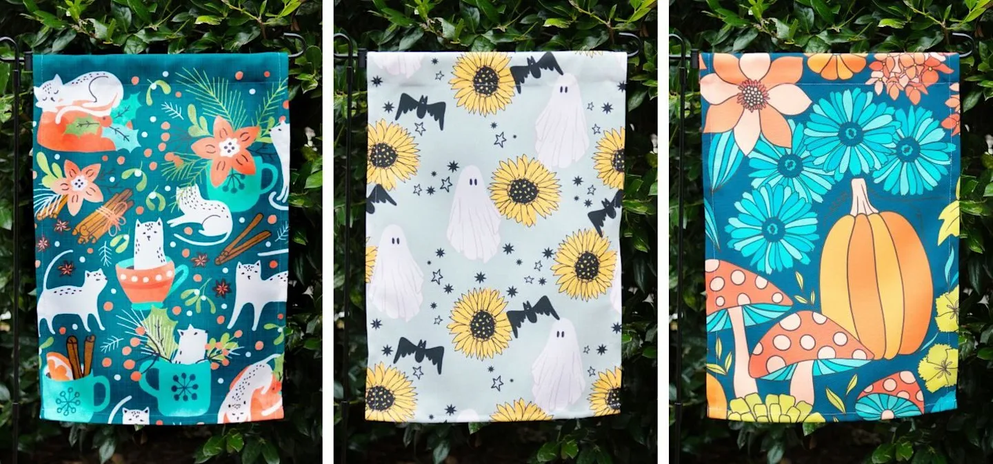 Three garden flags against a dark green bush: The first flag has white cats, green leaves and berries. Second has white ghosts and yellow sunflowers, third has a big pumpkin, some mushrooms and flowers.