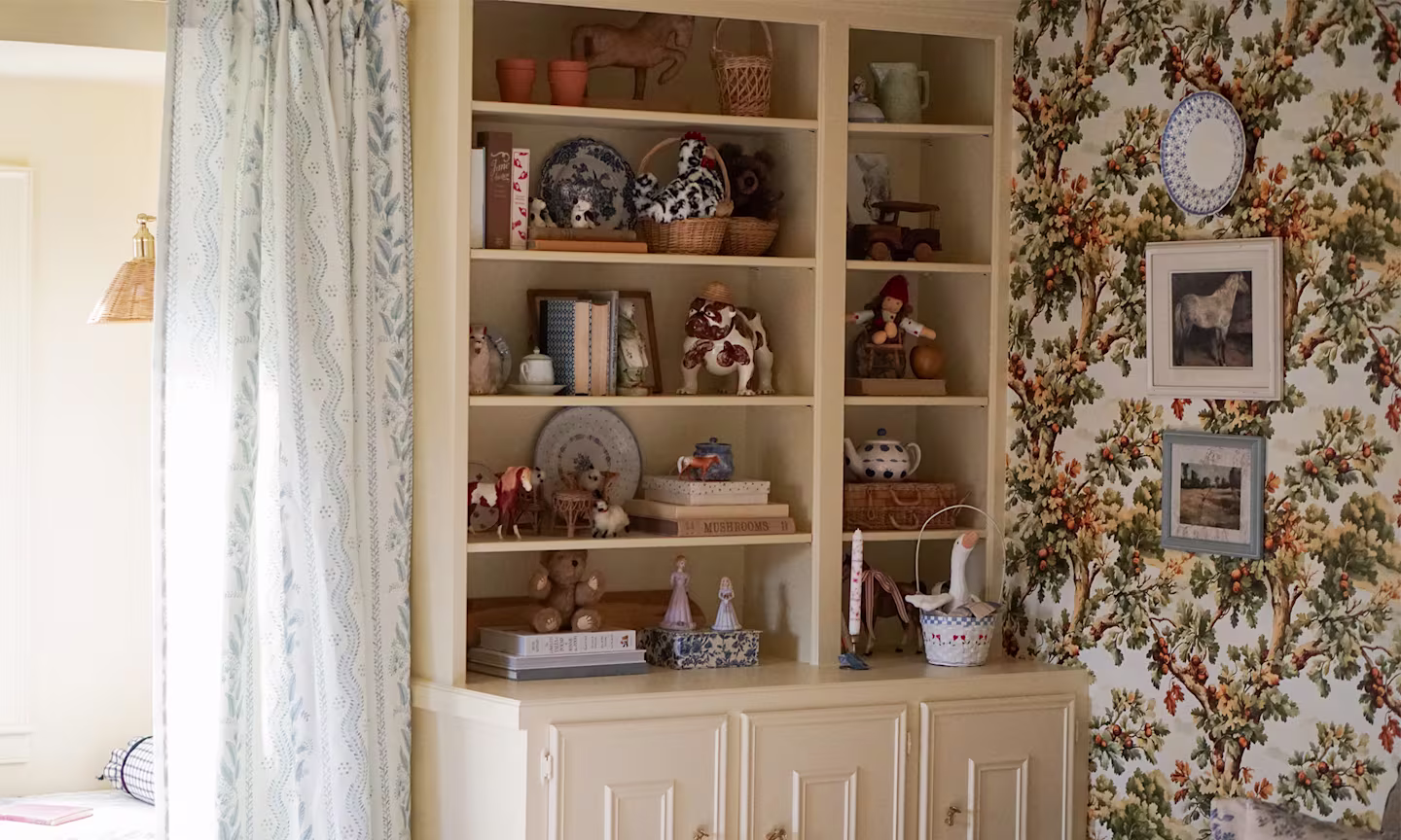 Playroom with oak tree wallpaper, open shelving and blue floral stripe curtains.