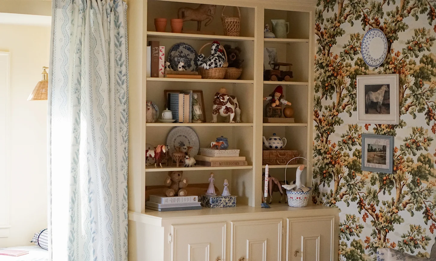 Playroom with oak tree wallpaper, open shelving and blue floral stripe curtains.