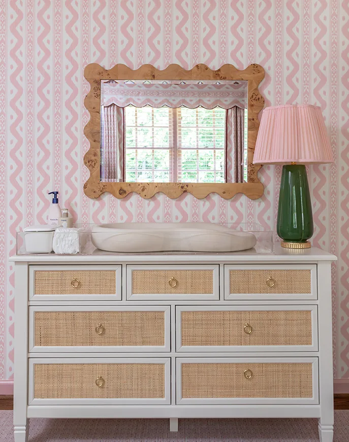 White dresser with cane-front drawers beneath a scalloped wood mirror, set against pink patterned wallpaper.