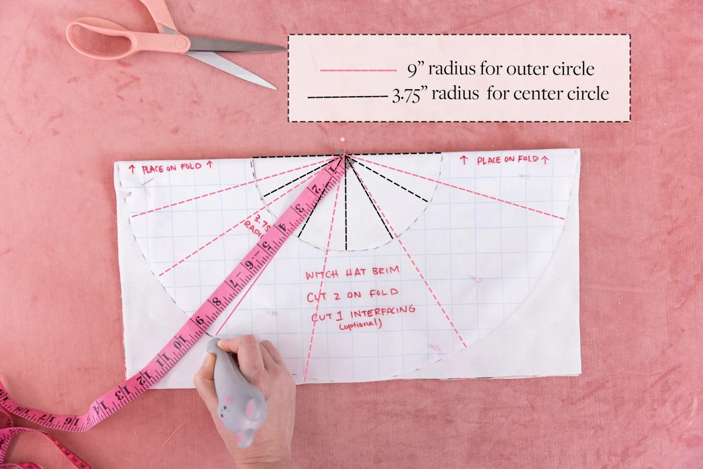 A circle template on grid paper is pinned down on top of a piece of folded fabric, with the design side in. A pink measuring tape is pinned at the top center of the folded fabric, extending out, to indicate how to measure the hat’s brim. Black and red das