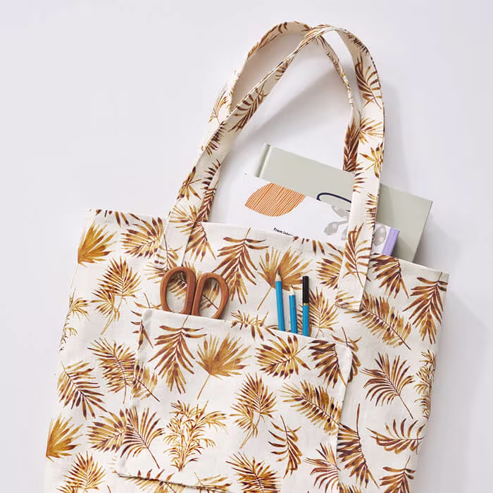 Fabric tote bag with golden-brown leaf pattern, shown with art supplies and notebooks tucked into the front pocket.