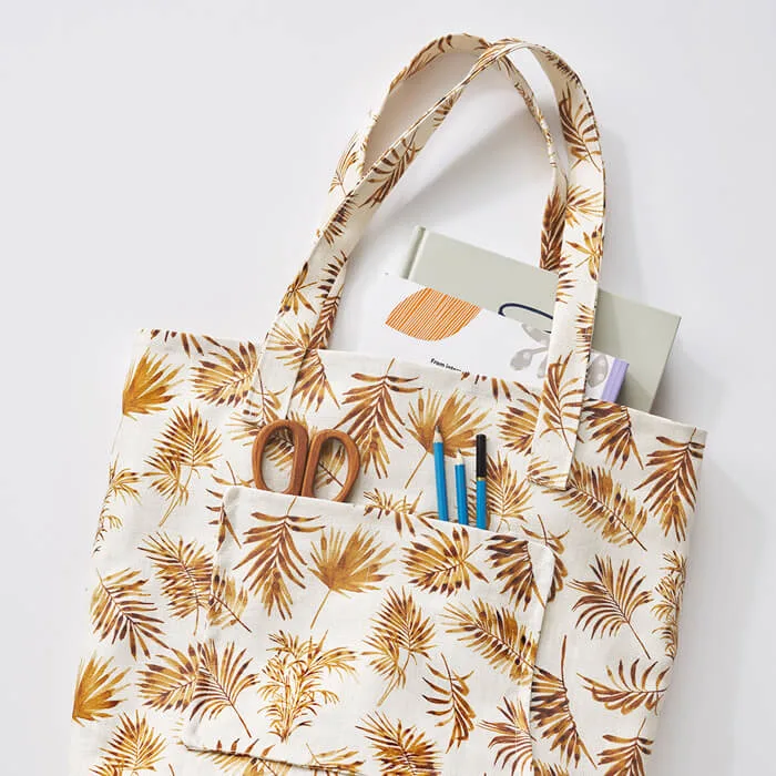 Fabric tote bag with golden-brown leaf pattern, shown with art supplies and notebooks tucked into the front pocket.