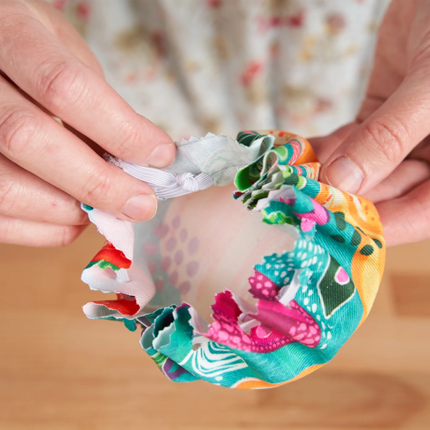A person pulling back the edge of fabric showing where two ends of white elastic have been tied together where they meet after being woven around the edge of a piece of fabric with holes punched near the edge. The design on the fabric has a teal backgroun