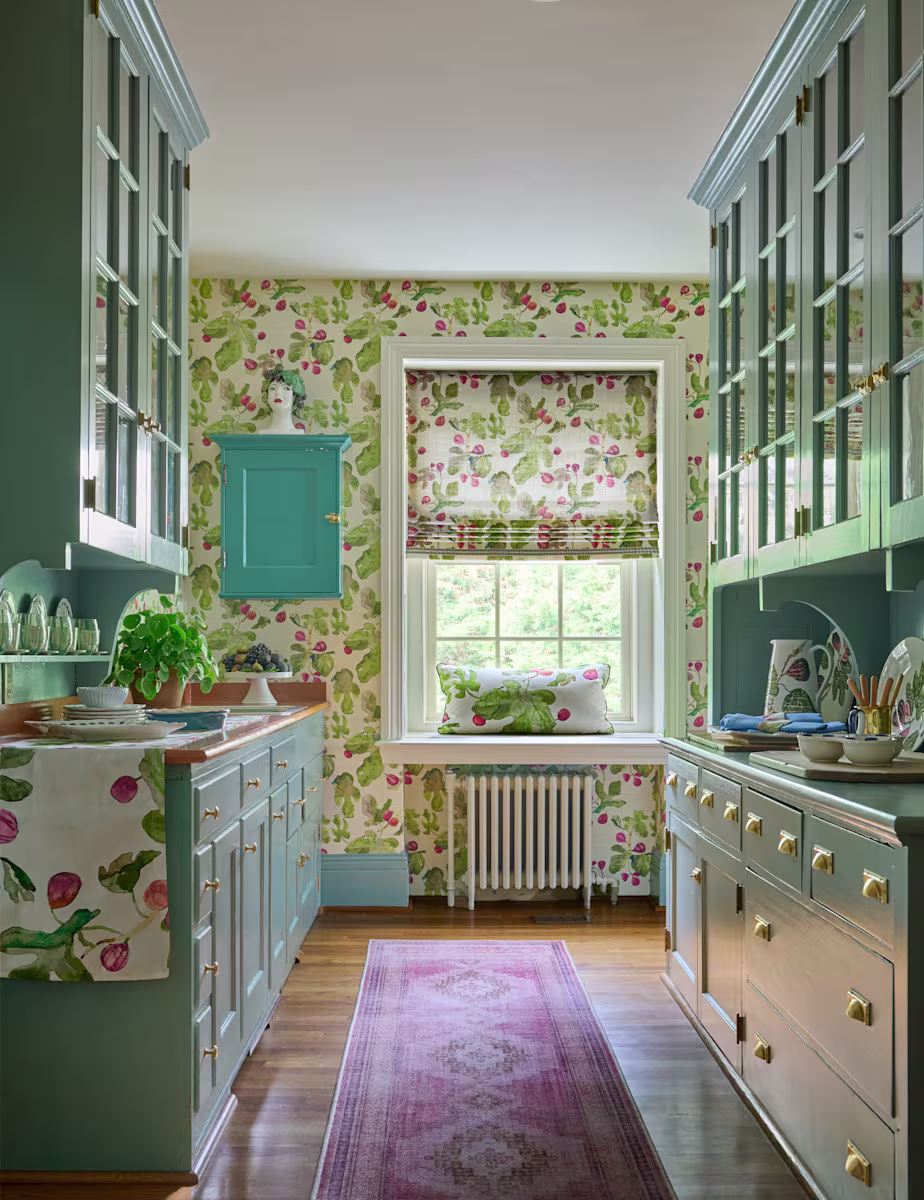 Traditional galley kitchen featuring botanical fruit wallpaper, coordinating Roman shade, sage-green cabinetry with brass hardware, wood flooring, and colorful decorative accents.