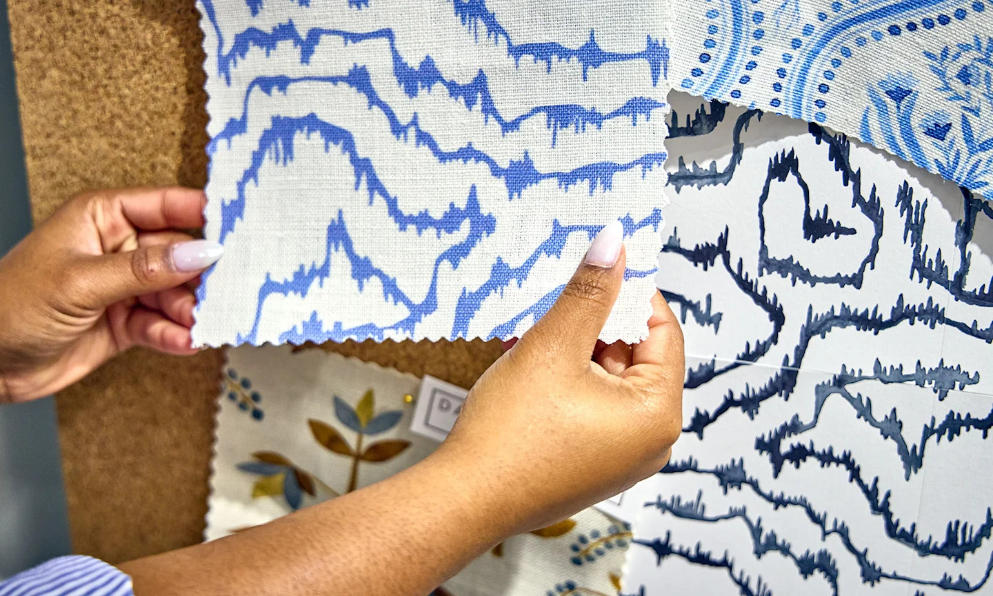 Hands holding swatches of fabric in a designs that has blue abstract lines on a cream background.