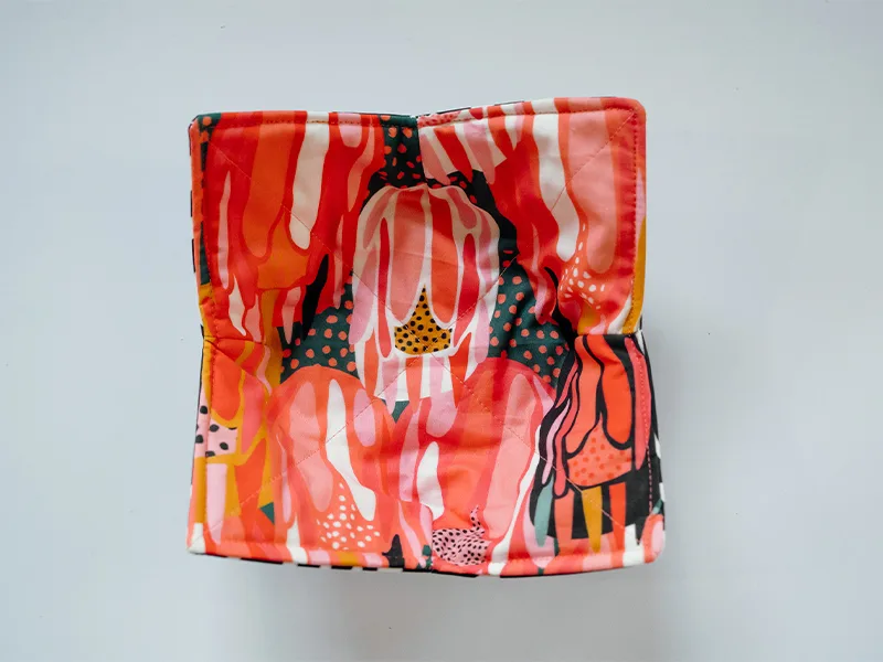 The finished bowl cozy as seen from above on a white surface. It has a concave shape to fit a bowl of soup and four pointed edges. The fabric on the top side features bright flowers in inks, reds, yellows and white. The fabric on the bottom side features 