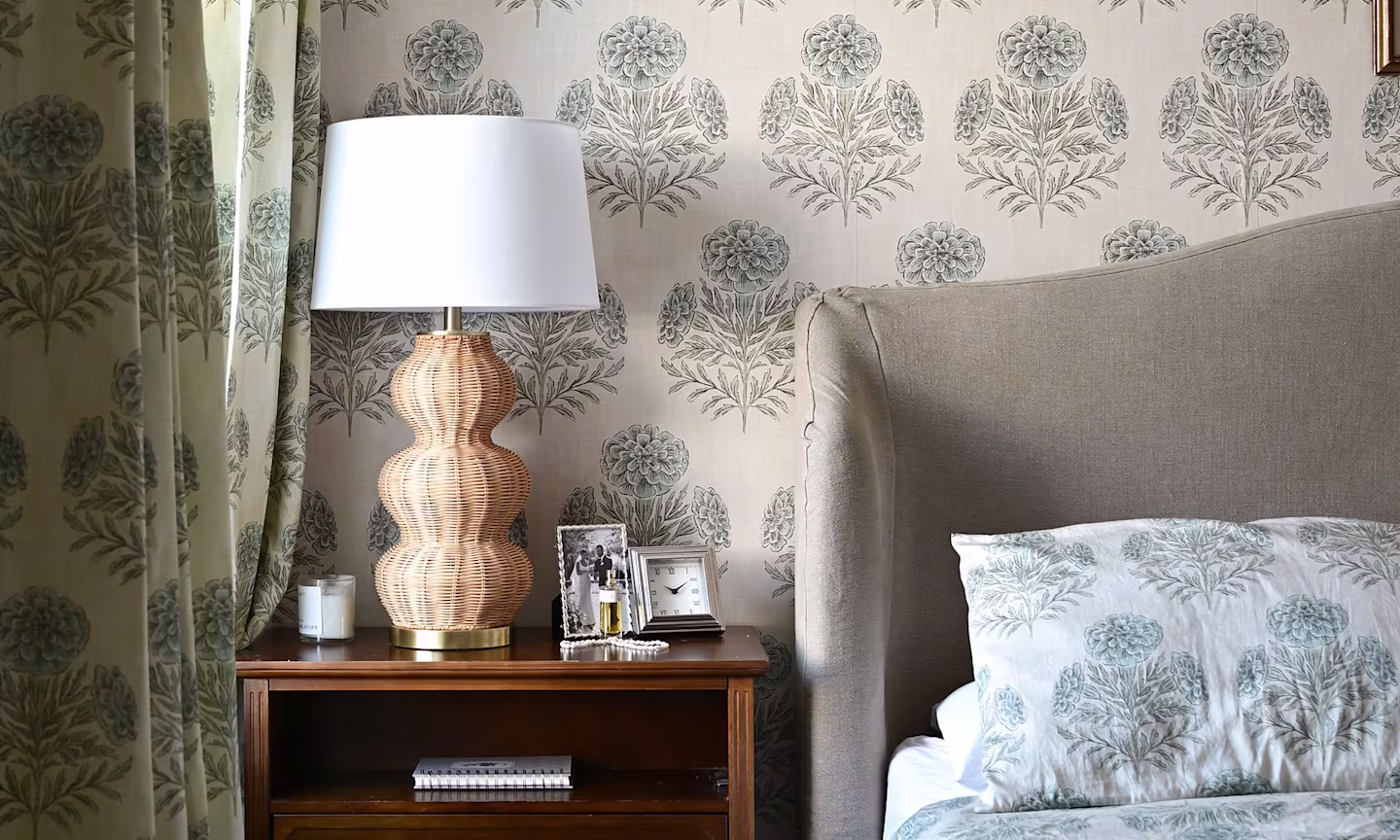 A cozy, elegant bedroom featuring soft blue-gray floral wallpaper and matching bedding. A woven wicker table lamp sits atop a dark wood nightstand, complemented by framed décor and a candle.