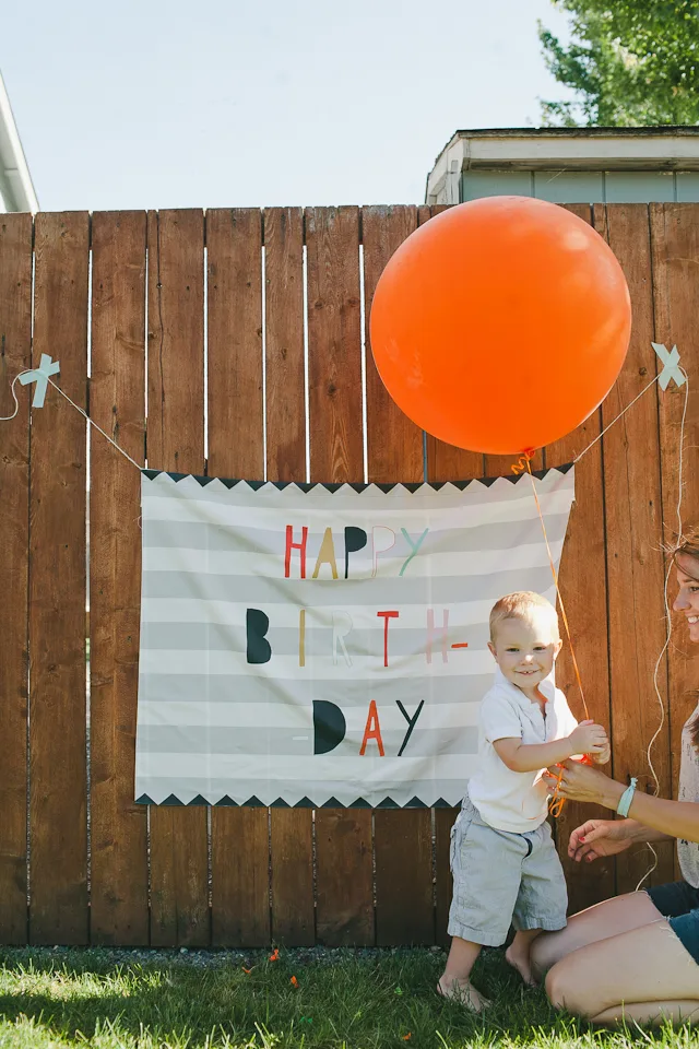 A small child stands in front of a fabric sign hung up against a wooden fence. The sign says "Happy birthday," A large orange balloon hangs to the top right of the sign.