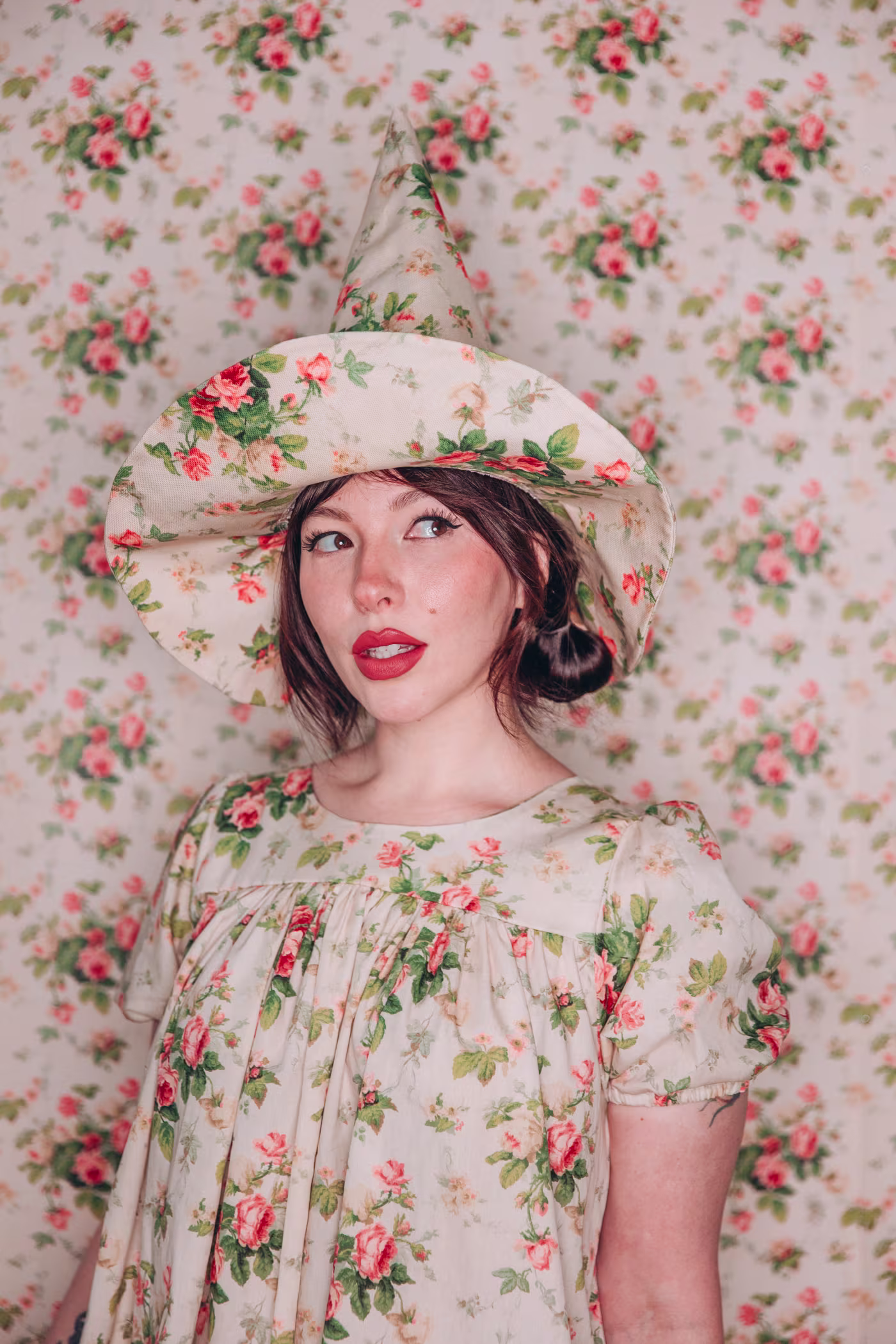 A close up of Keiko in a floral witch hat and matching dress, in front of a backdrop of the same design. She's looking to her right.