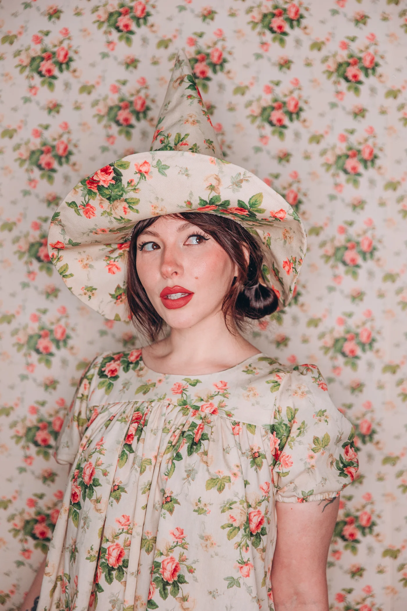 A close up of Keiko in a floral witch hat and matching dress, in front of a backdrop of the same design. She's looking to her right.