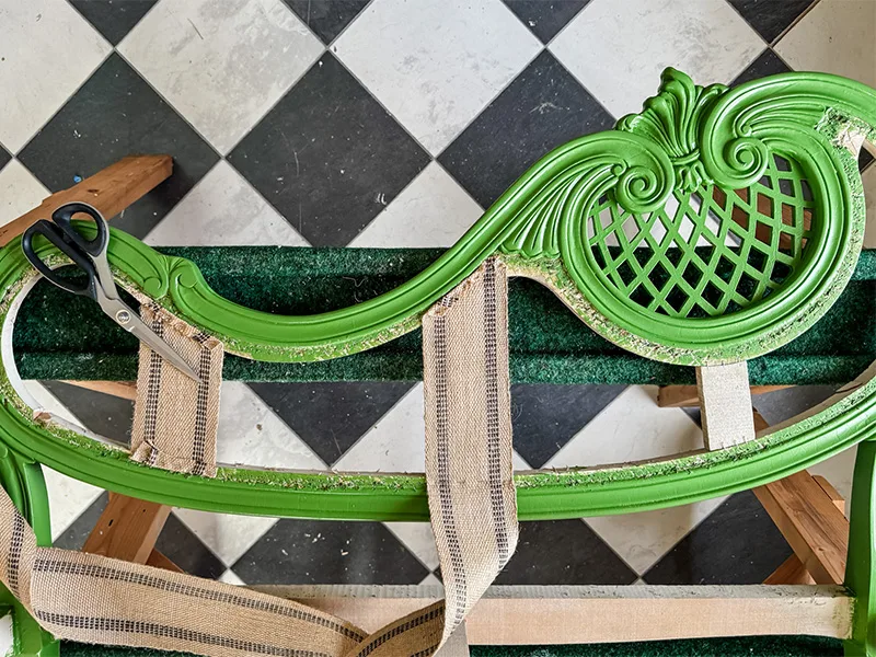 A close up of the settee top on a sawhorse with jute webbing stapled to the top. A pair of scissors sit near the edge. A checkered floor is in the background.