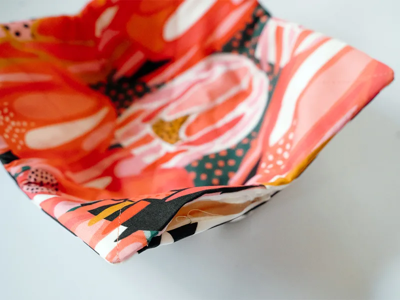 The bowl cozy has been turned inside out using a turning tool so that the fabric designs are visible on both sides. The fabric has been pulled through the 3” opening, which is visible. The fabric on the top side features bright flowers in pinks, reds, yel