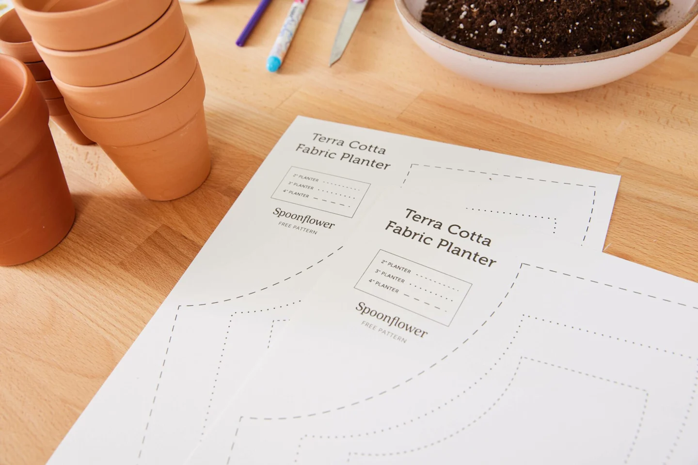 The printed template for the fabric-covered flower pot has been printed out twice, to make two fabric-covered flower pots. The paper pieces are lying on a wooden table with two stacks of terracotta pots to the left and a bowl of dirt to the top.