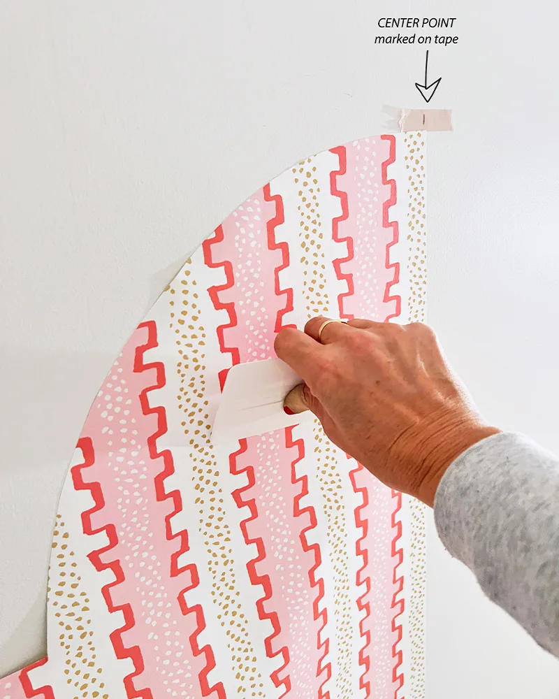 Installing the left half of the wallpaper headboard with a small white squeegee. A small pink piece of tape has been added to the center top of the headboard. A black arrow and the words "Center point, marked on tape" have been added and point to the piec
