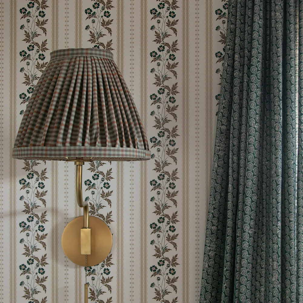 Close up of a wall with striped floral wallpaper, block printed curtains and a plaid fabric lampshade.