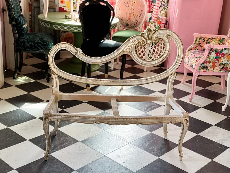 A settee chair stripeed down to the wooden frame on a checkered floor with other upholstered furniture behind it.