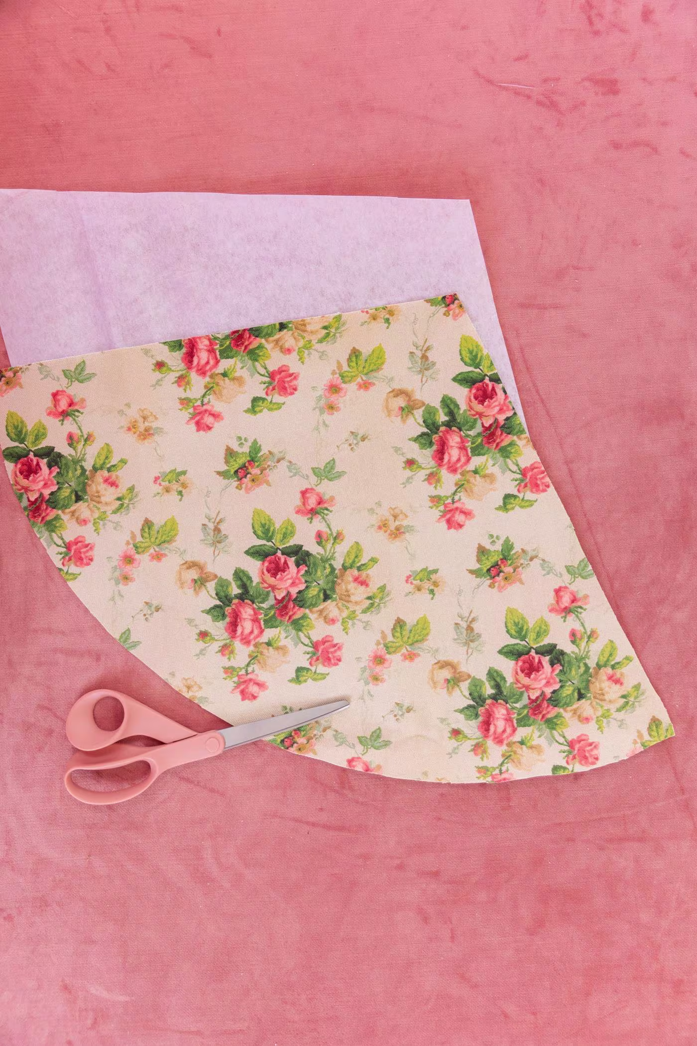 A pair of peach scissors rests on a piece of cone-shaped fabric with pink roses and green leaves behind a creme background. The heavy interfacing is behind the fabric. Pink carpet surrounds as the background.
