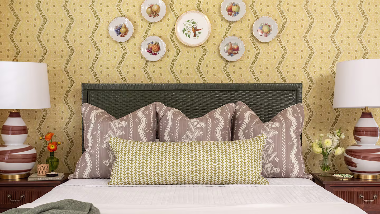Bedroom with yellow wallpaper, white linens, three throw pillow and one lumbar pillow.