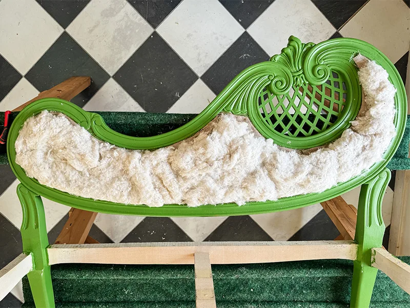 A layer of cotton on top of burlap on the frame of a green settee resting on a sawtable, with a checkered floor in the background.