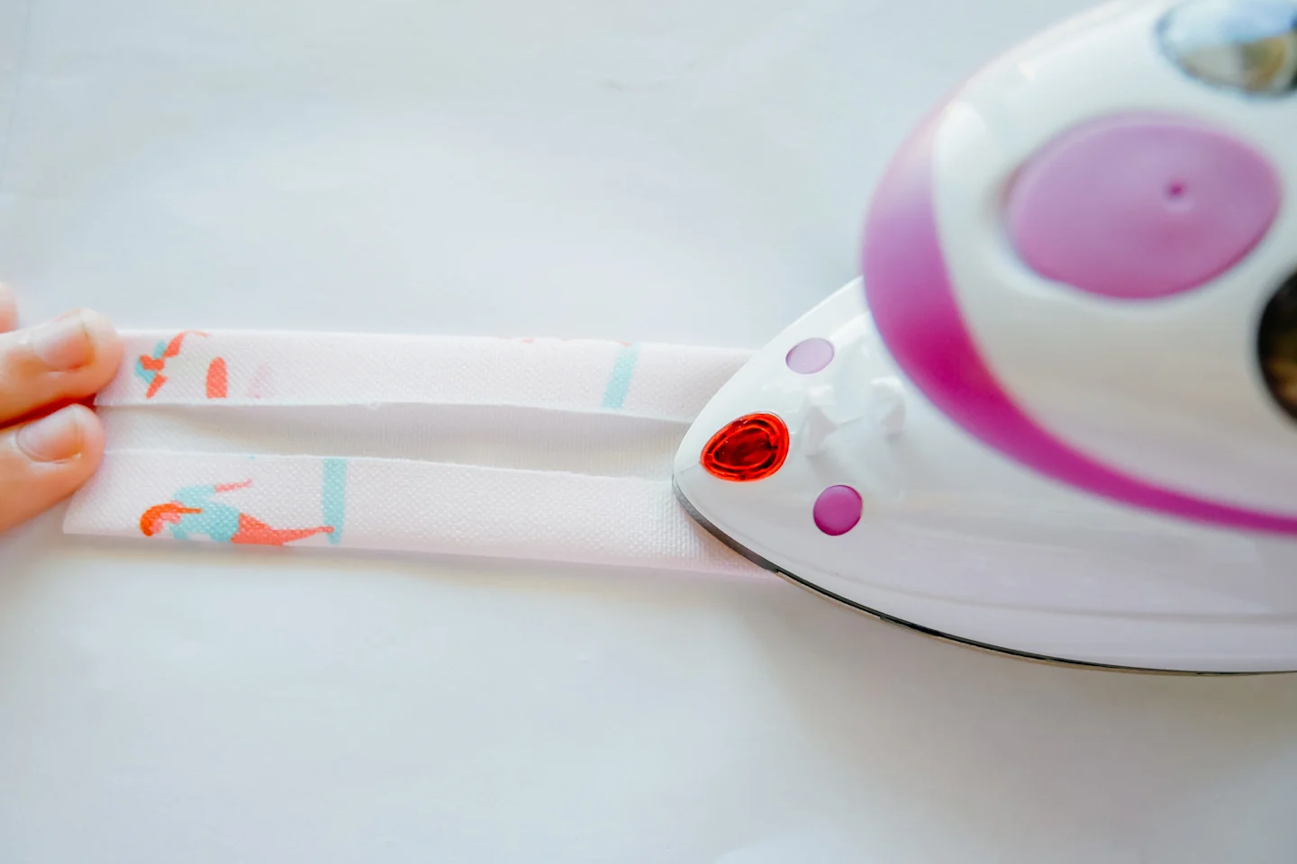 A small rectangle of fabric is being pressed lengthwise with a hot iron. The left edge of the fabric and the right edge of the fabric are being pressed toward the middle with a small gap between the two. The fabric features a design with small female surf
