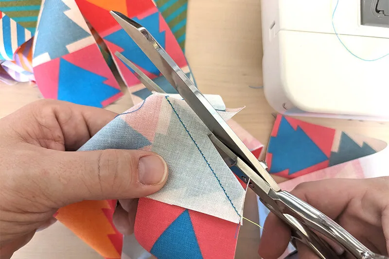 Lisa is cutting extra fabric left over from when two top corners of perpendicular strips of fabric were sewn together at a 45-degree angle. She cutting off the small extra triangles of fabric that are no longer needed.