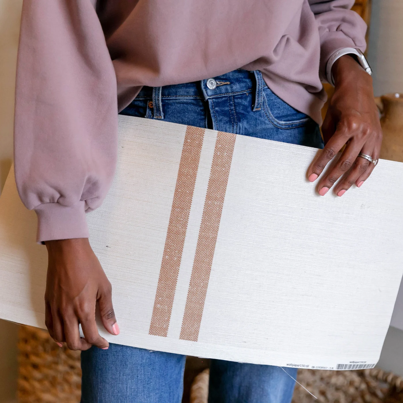 A photo of Tiffany holding a swatch of grasscloth wallpaper down by her waist. Her hands, torso and legs are shown. She is wearing a mauve blouse tucked into blue jeans. The design on the wallpaper has a cream background and two small dark tan repeating s