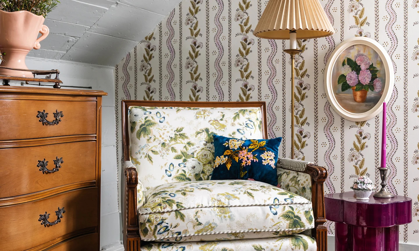 Corner of a guest bedroom with purple floral stripe wallpaper and a green chintz upholstered chair.