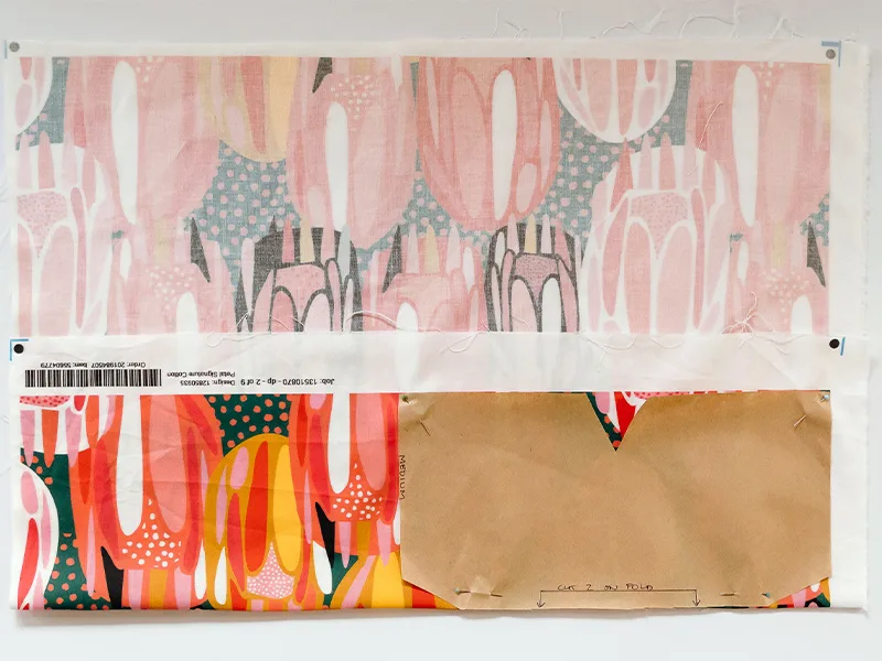 The bowl cozy paper pattern is pinned and placed on the fold of a piece of fabric and ready for cutting out. The fabric design features bright flowers in pinks, reds, yellows and white.