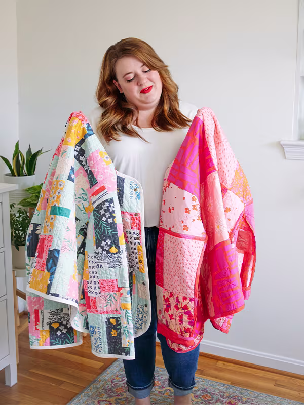 Meg holds up two cheater quilt quilted jackets featuring delicate florals. The one on the left features prints with mostly with a blue or pink theme. The one on the right features mostly pink prints. She is standing in front of a white wall.