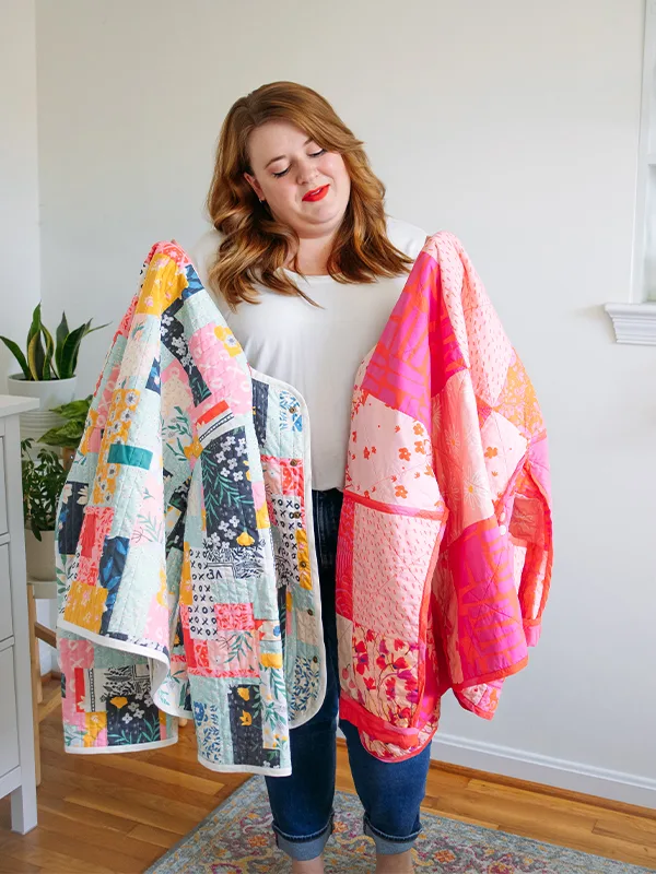 Meg holds up two cheater quilt quilted jackets featuring delicate florals. The one on the left features prints with mostly with a blue or pink theme. The one on the right features mostly pink prints. She is standing in front of a white wall.